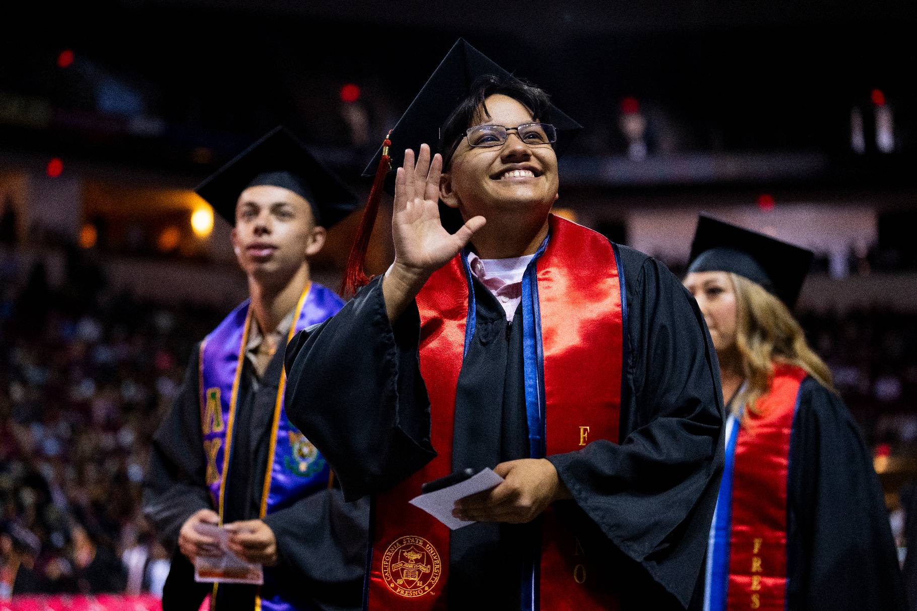 Male commencement student walking among his peers waving his hand to the crowd